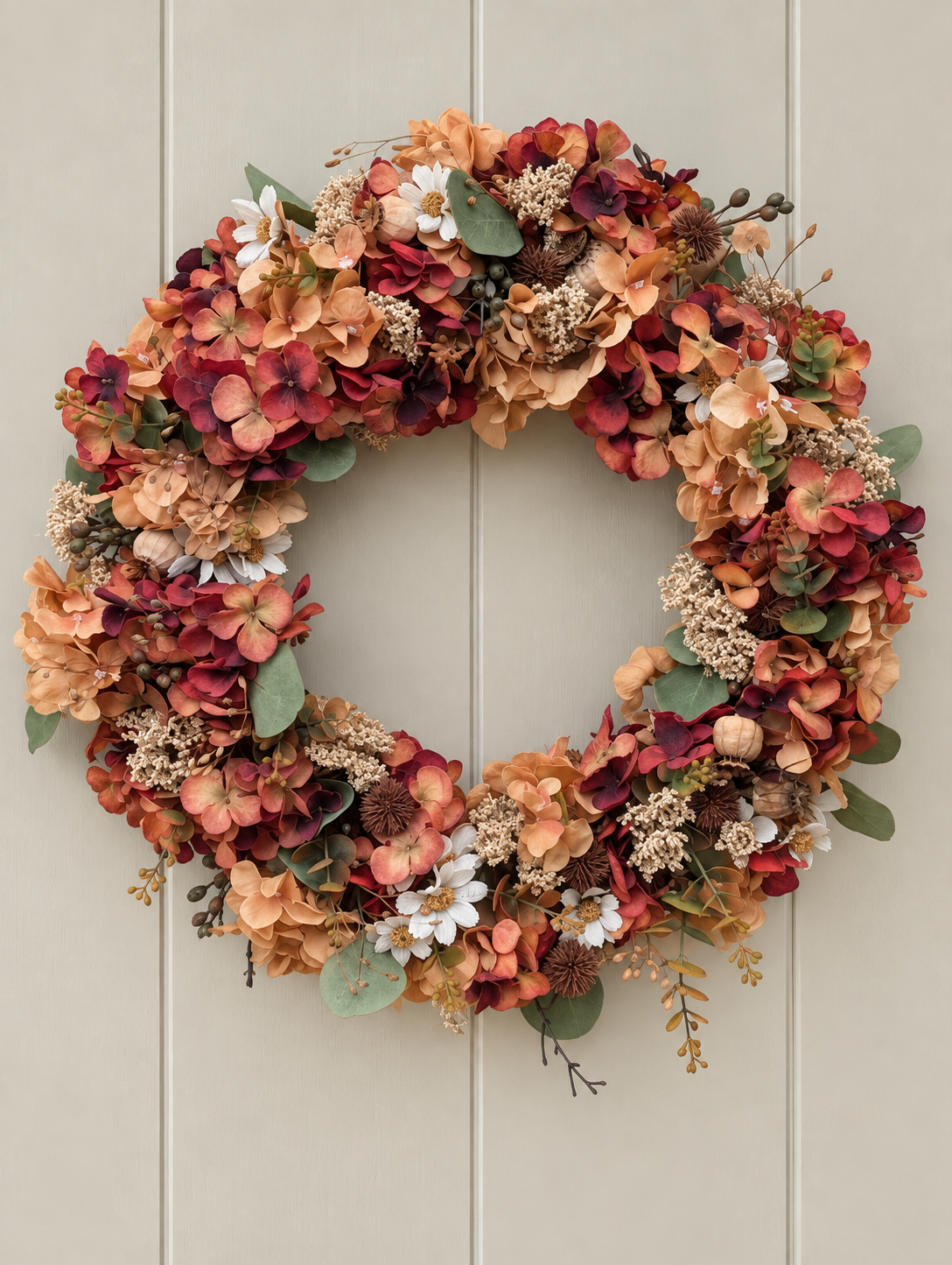 An autumn-themed floral wreath featuring hydrangeas and dried flowers in shades of rust red, peach, orange, cream, and green, hung on a light beige paneled door.