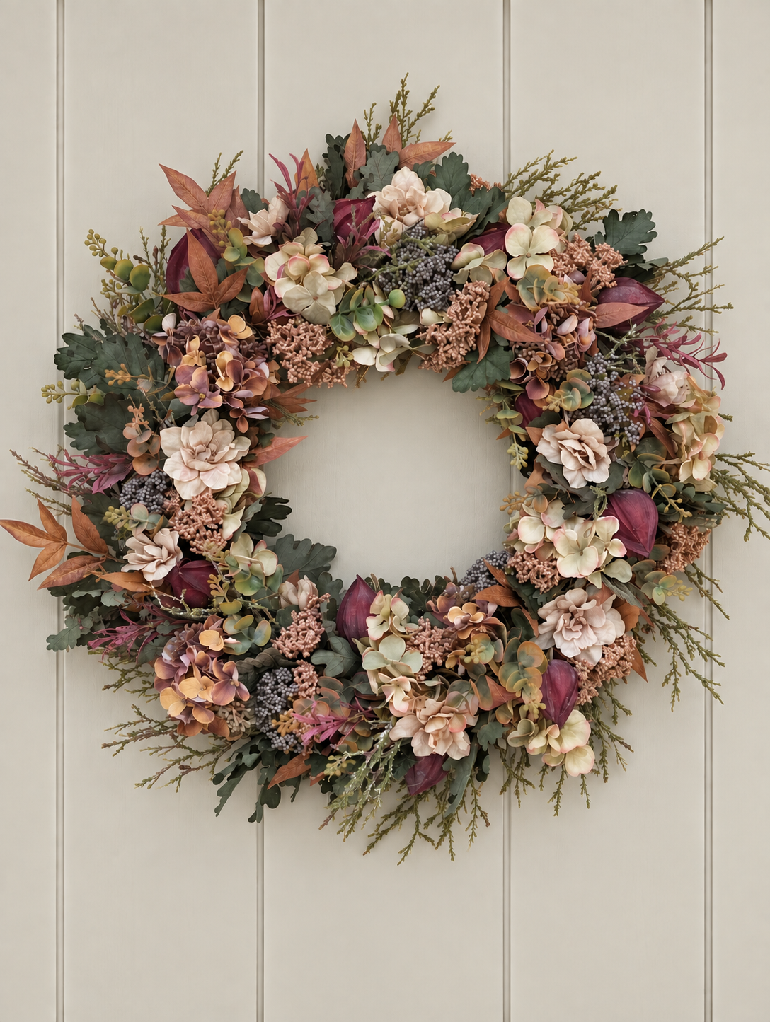 An autumn-themed floral wreath with cream, burgundy, and tan flowers, green and brown leaves, and dried botanicals, hanging on a light beige paneled door.