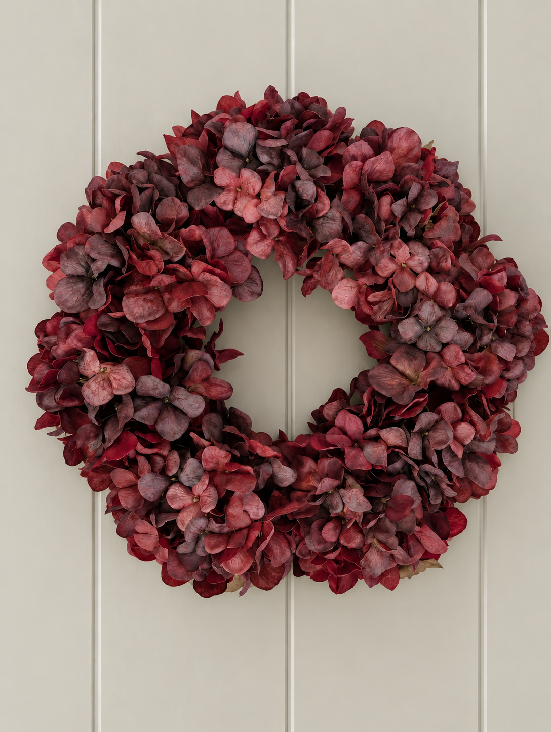 A circular faux hydrangea wreath in deep reds, burgundy, and dusty pink tones, hung against a light beige paneled door.