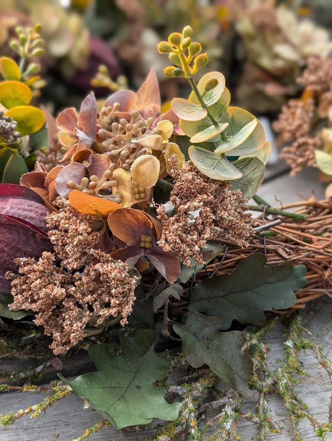 An autumn-themed artificial floral arrangement featuring dried brown and orange hydrangea-like blooms, green eucalyptus leaves, and brown grapevine stems on a weathered gray wood surface.