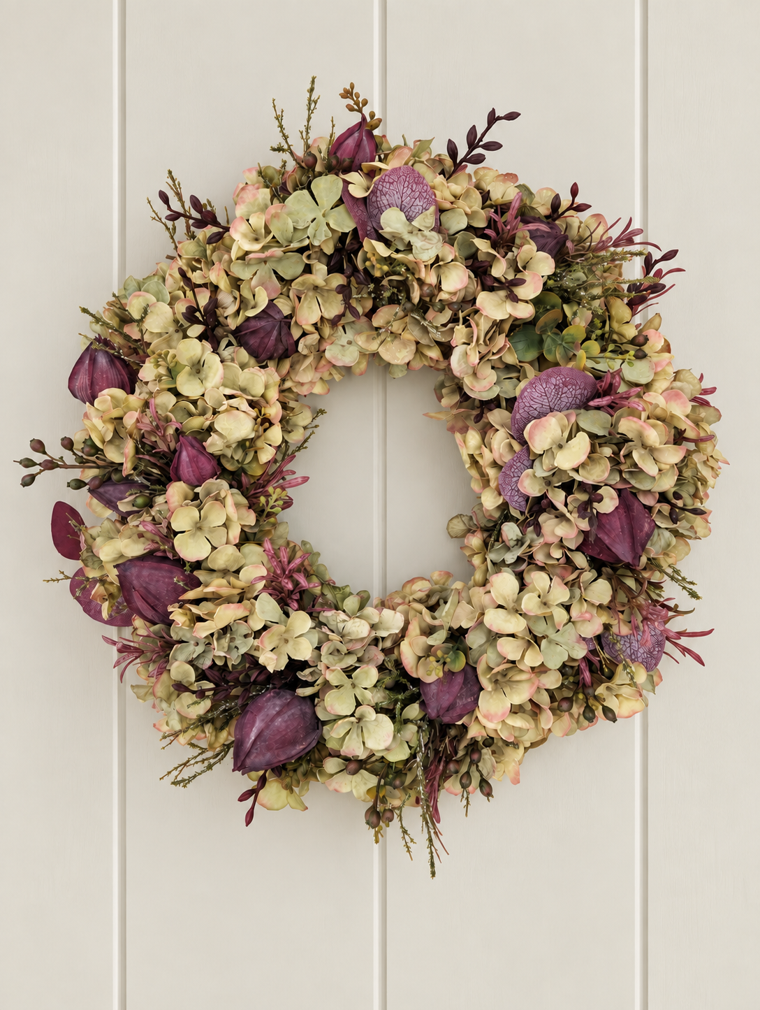 A round artificial floral wreath with dried hydrangea-like cream flowers, muted green leaves, and prominent deep purple accents, hanging against a white paneled door.