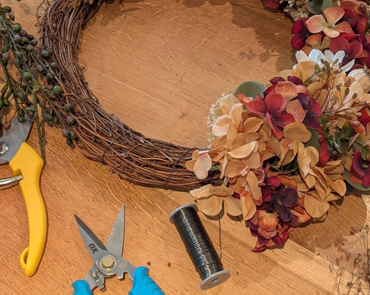 A rustic floral wreath made of brown grapevine branches, filled with dried beige, burgundy, and white flowers, green leaves, and eucalyptus stems, displayed on a wooden table with floral shears and a spool of wire. Nationwide delivery.